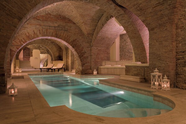 An indoor swimming pool with arched brick ceilings, lounge chairs, and decorative lanterns.
