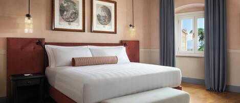 Un dormitorio con una cama grande, un banco, una mesita de noche, dos cuadros enmarcados y una ventana con cortinas.
