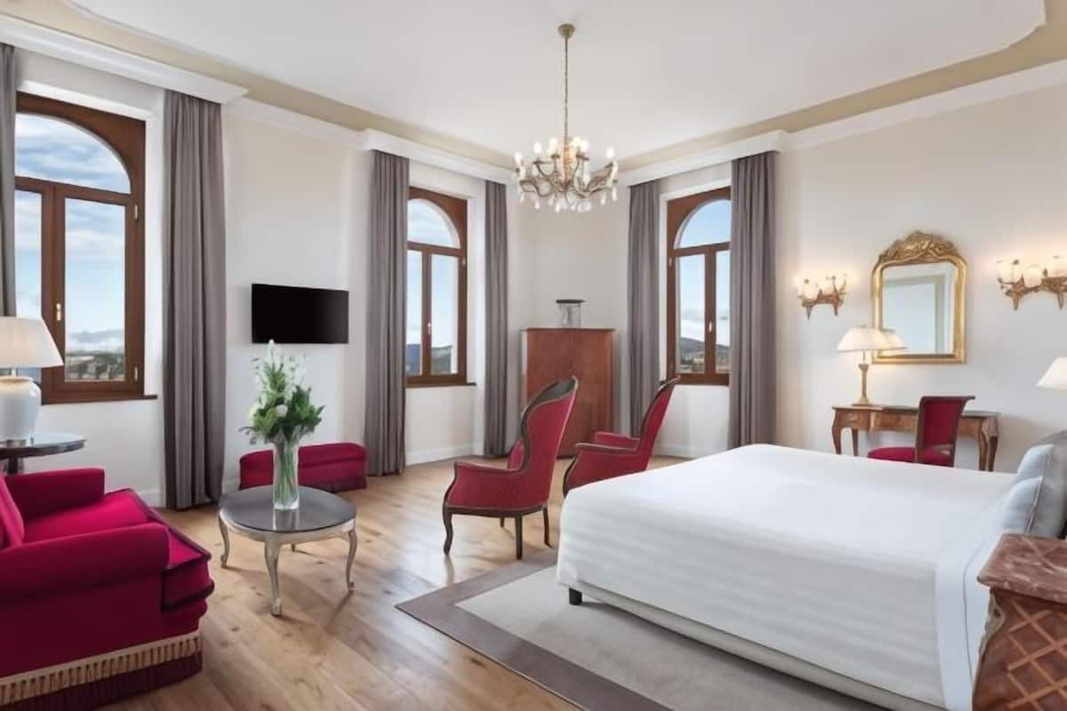 A hotel room with a large bed, a desk with a chair, a TV, and a chandelier.