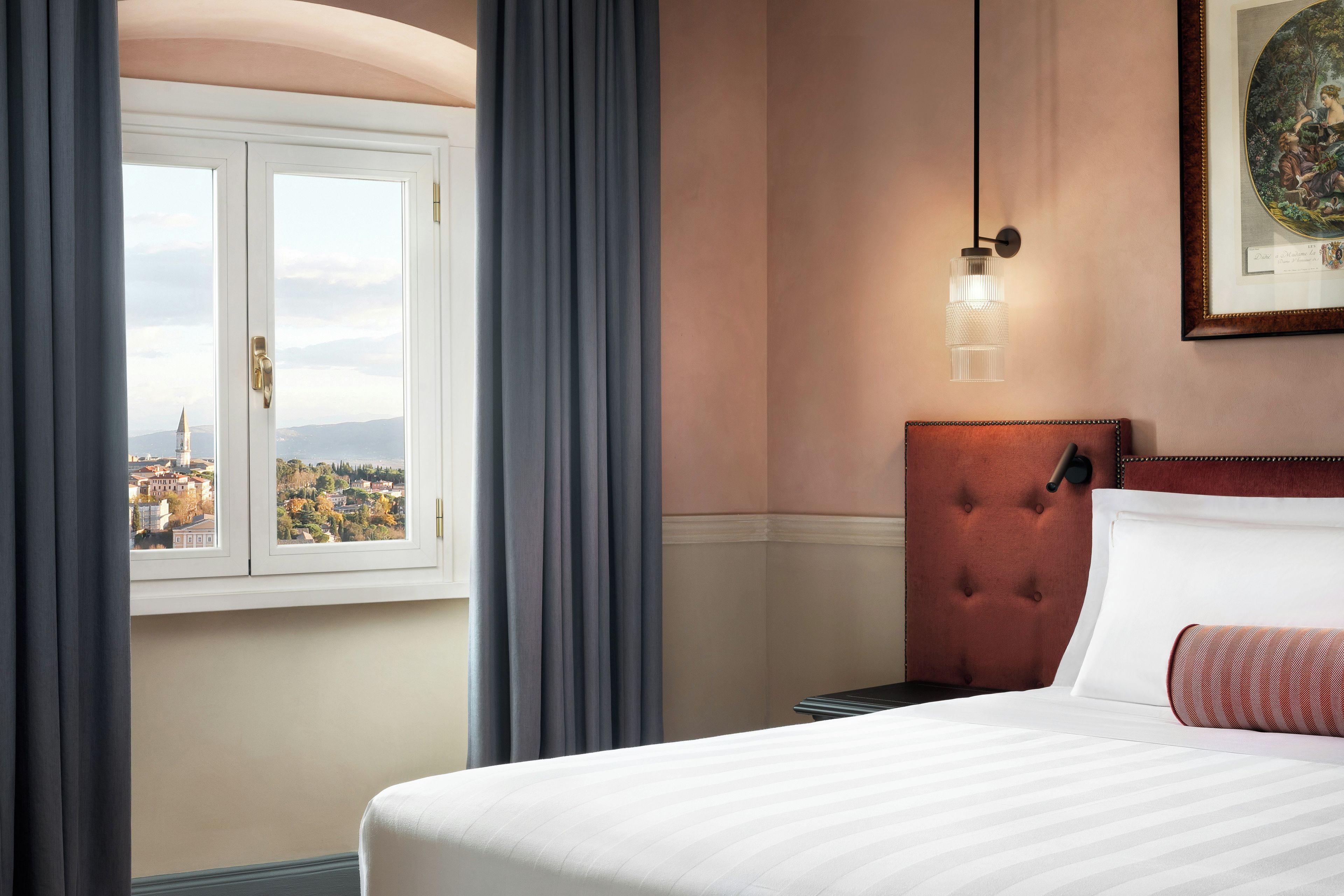 Une chambre d’hôtel avec un lit, une vue par la fenêtre, une tête de lit capitonnée et un tableau encadré au mur.