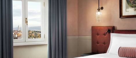 A hotel room with a bed, a view through a window, a tufted headboard, and a framed picture on the wall.