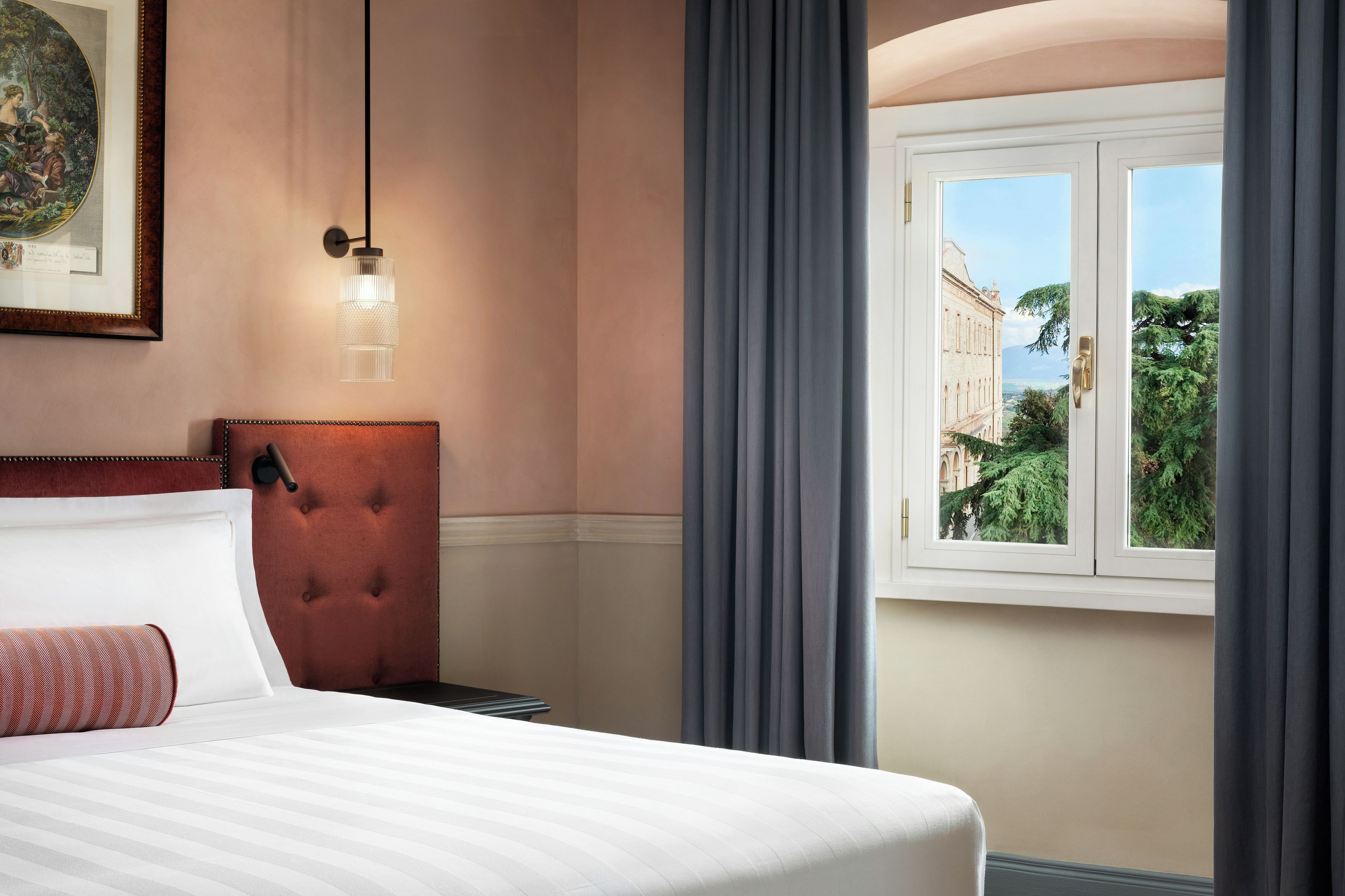 A hotel room with a bed, a tufted headboard, a framed picture, a hanging light fixture, and a window with curtains.
