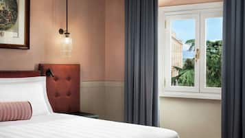 Une chambre d’hôtel avec un lit, une tête de lit capitonnée, un tableau encadré, un luminaire suspendu et une fenêtre avec des rideaux.