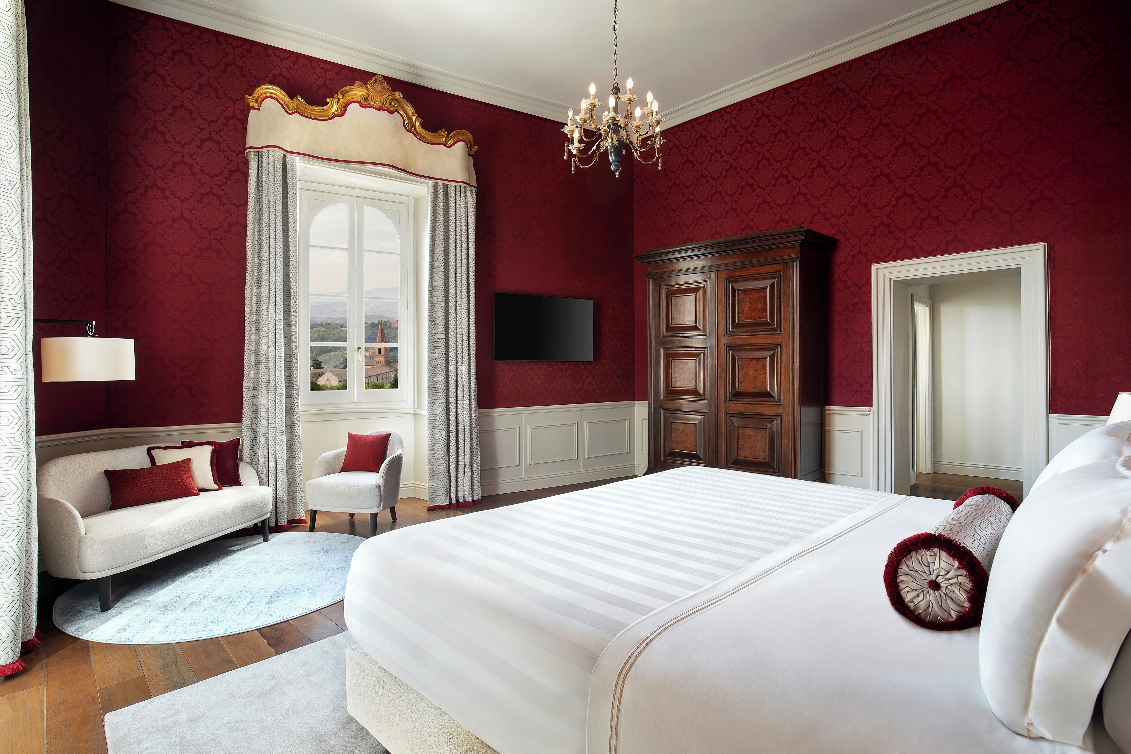 A hotel room with a large bed, a sofa, a chair, a wooden wardrobe, a TV on the wall, and a chandelier.