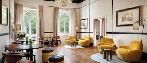 A spacious room with a chandelier, wooden flooring, yellow armchairs, a round table, and large windows with curtains.