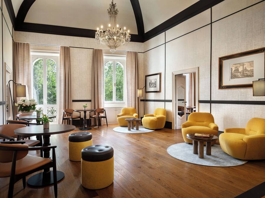 Amplia habitación con candelabro, suelo de madera, sillones amarillos, una mesa redonda y ventanas grandes con cortinas.