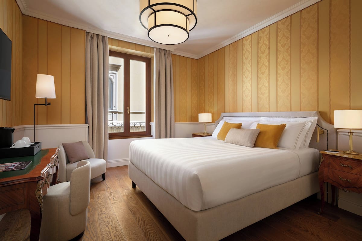 A hotel room with a large bed, a desk with a chair, a lamp, and a window with curtains.