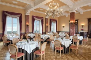A dining area with round tables, wicker chairs, chandeliers, and large windows with curtains. - Sina Brufani (Perugia)
