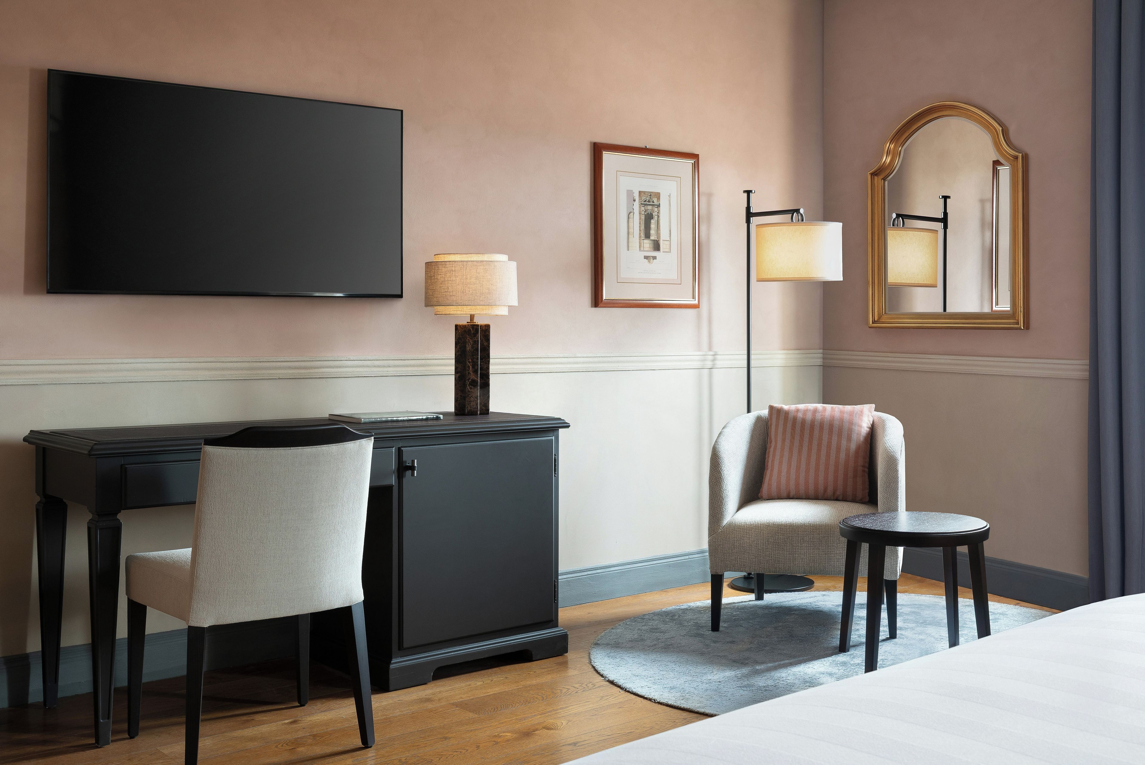 A hotel room with a flat-screen TV, a desk with a chair, a round side table, a sofa, and a large mirror.