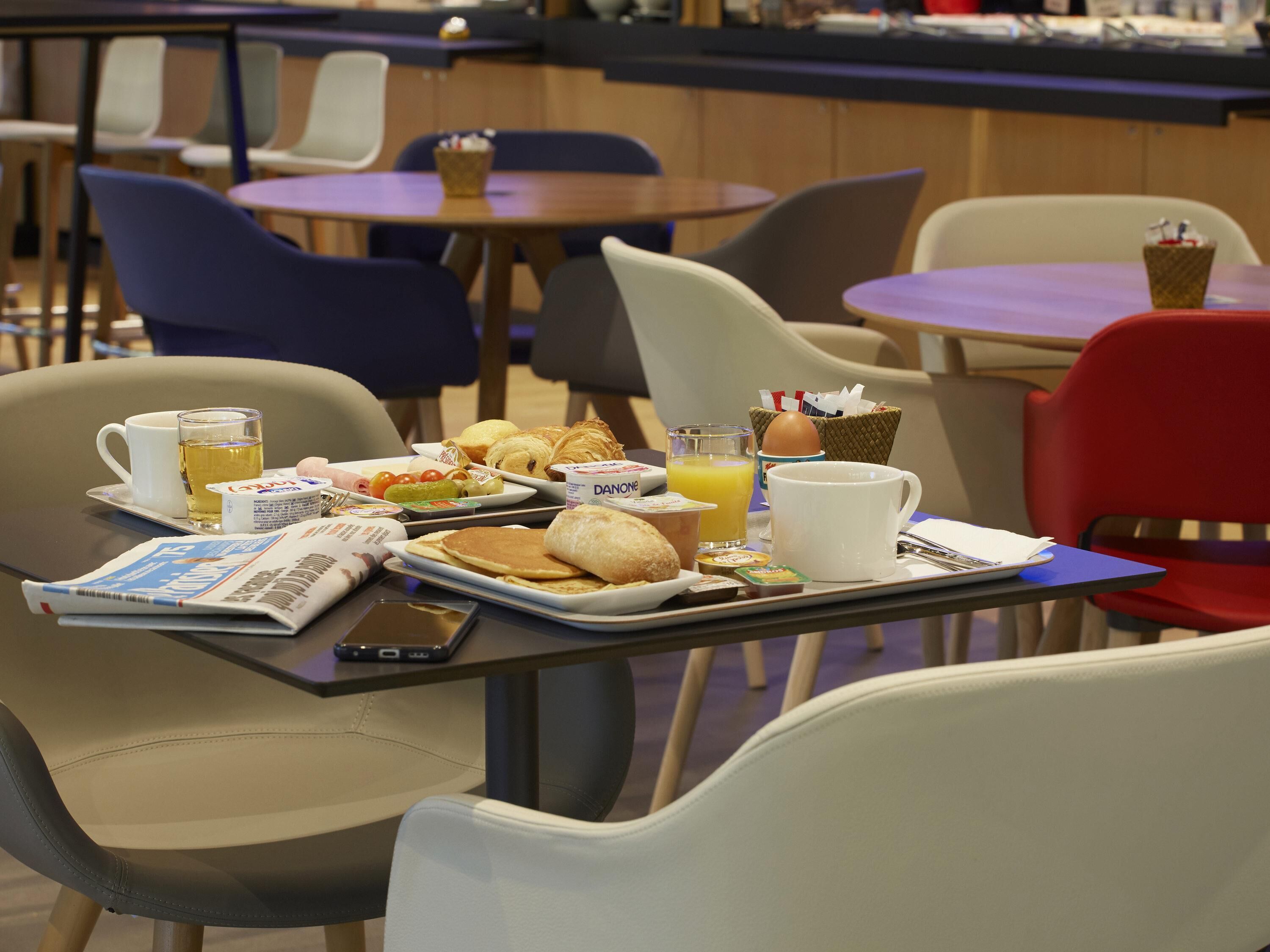 Una bandeja de desayuno con diversos alimentos, un periódico y un vaso de jugo de naranja sobre una mesa en un comedor con sillas y mesas.