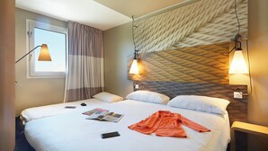 A hotel room with a large bed, two bedside tables, a window with curtains, and a textured wall with wall-mounted lamps. - ibis Archamps Porte de Geneve (Archamps)