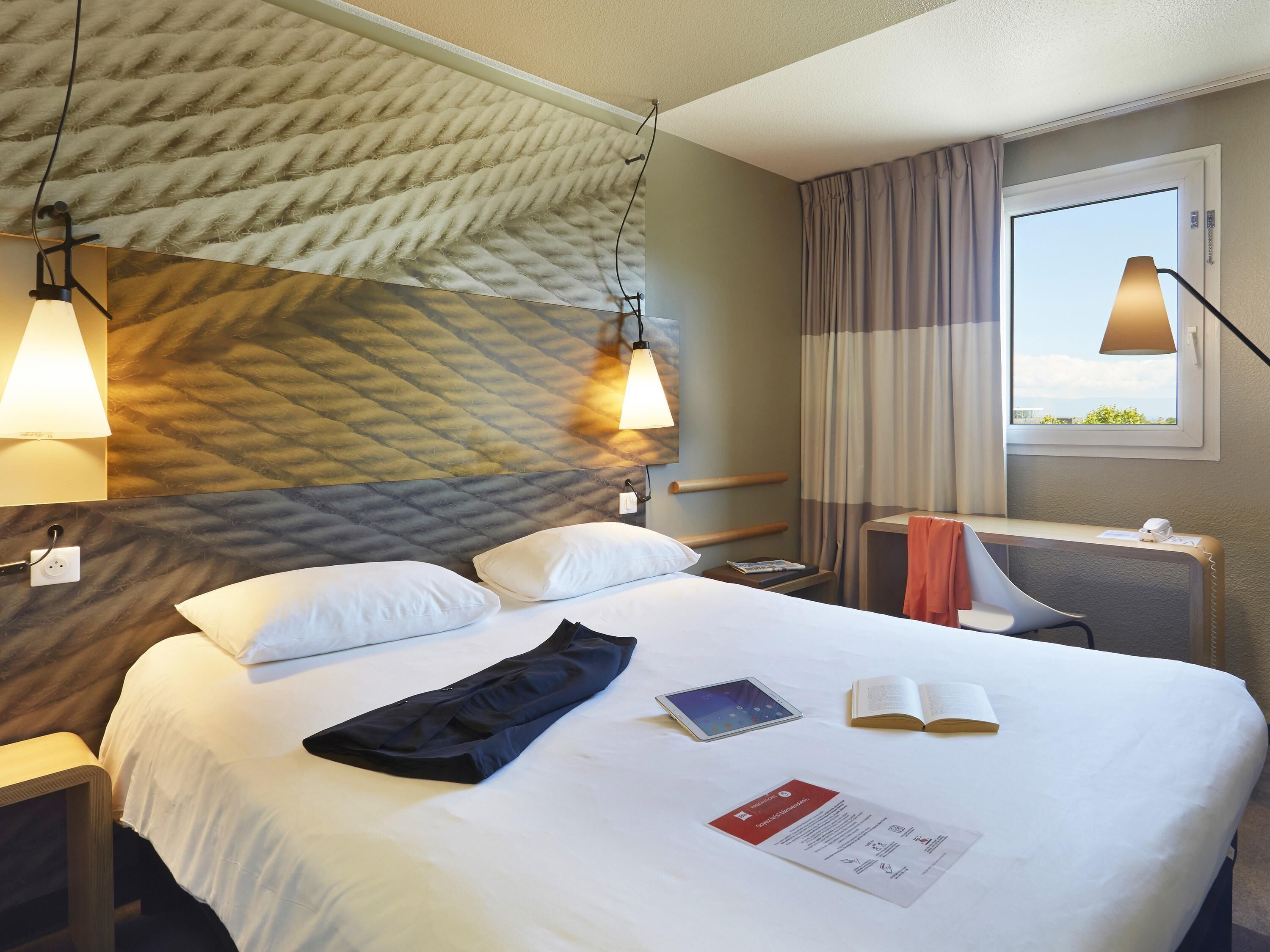 Een hotelkamer met een bed, nachtlampjes, een nachtkastje, een stoel, een raam en uitzicht op de lucht.