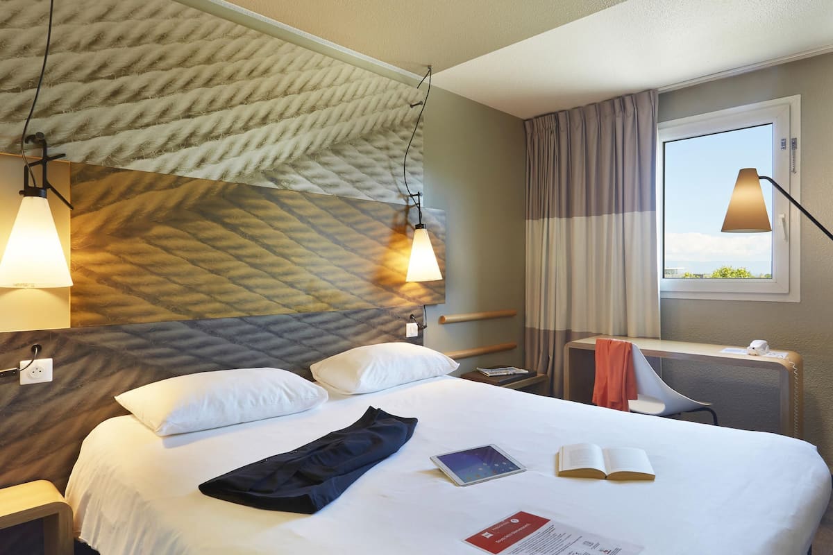 A hotel room with a bed, bedside lamps, a bedside table, a chair, a window, and a view of the sky.