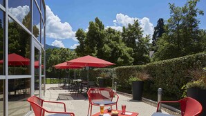 An outdoor seating area with red chairs and tables, surrounded by glass walls and a red umbrella. - ibis Archamps Porte de Geneve (Archamps)