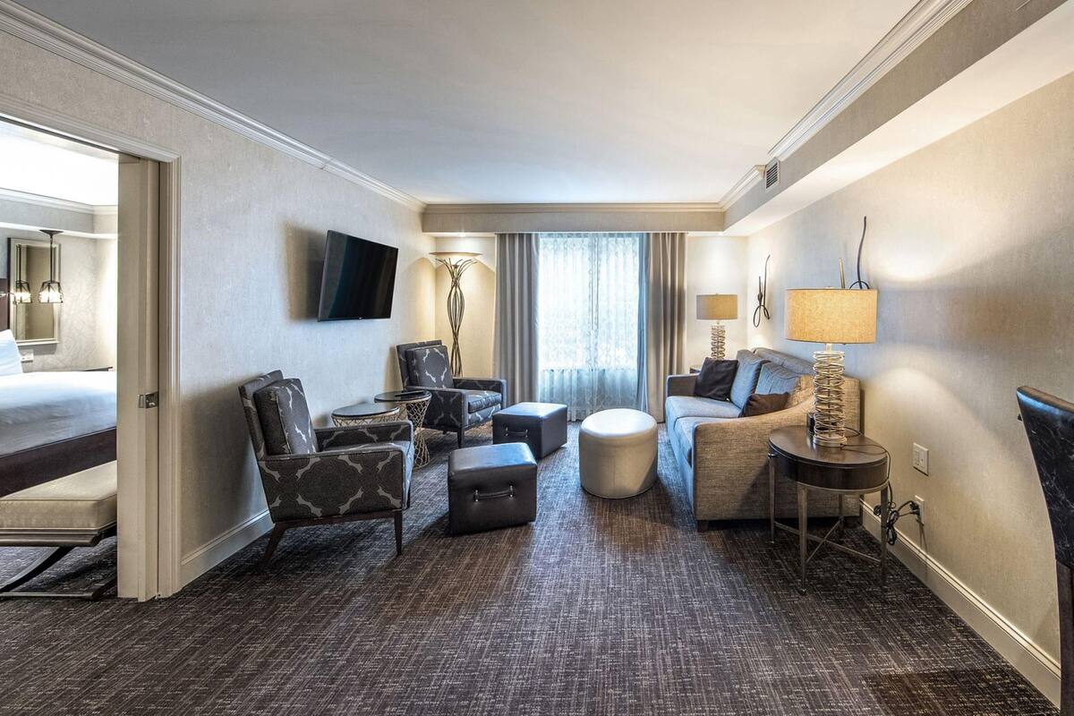 Una habitación de hotel con cama, zona de estar con sofá y sillones, una mesita y una lámpara.