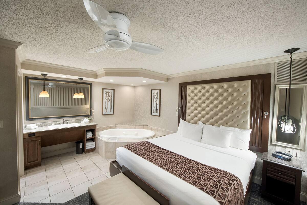 Habitación de hotel con una cama grande, baño independiente con bañera, tocador con espejo y ventilador de techo.