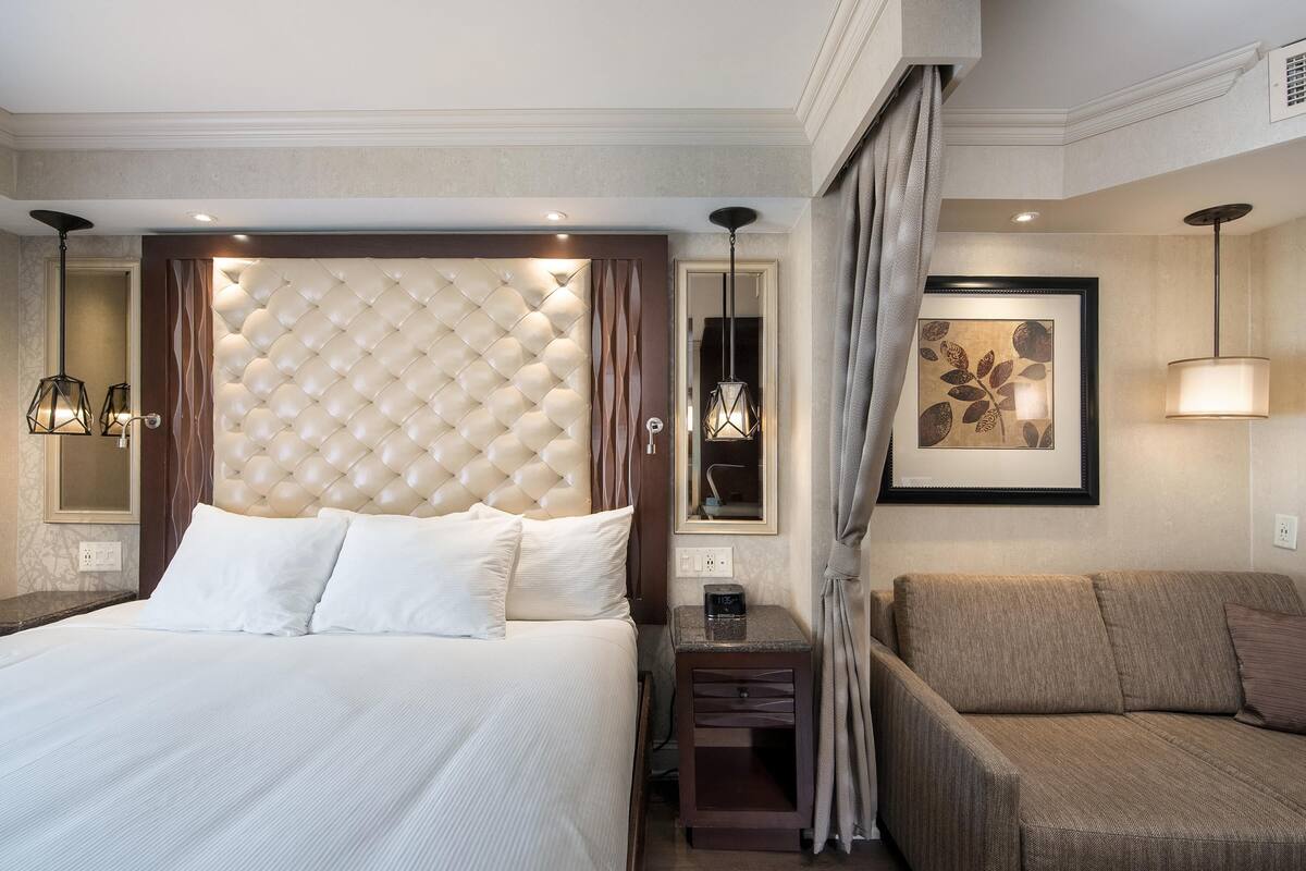 Habitación de hotel con una cama grande, un sofá, una mesita de noche con lámpara y una obra de arte enmarcada en la pared.