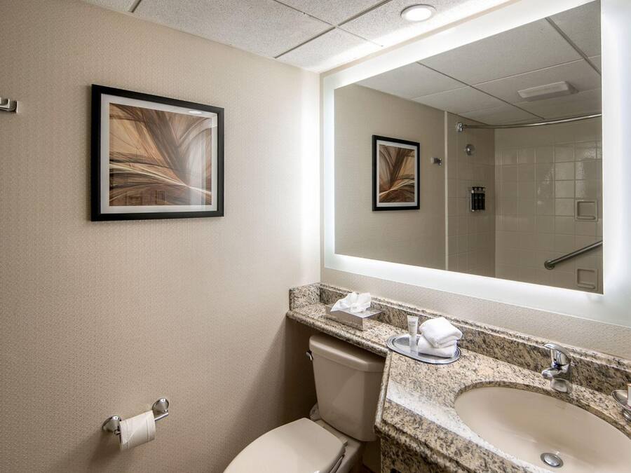 A hotel bathroom with a toilet, sink, and mirror. There are framed pictures on the wall and a towel dispenser.