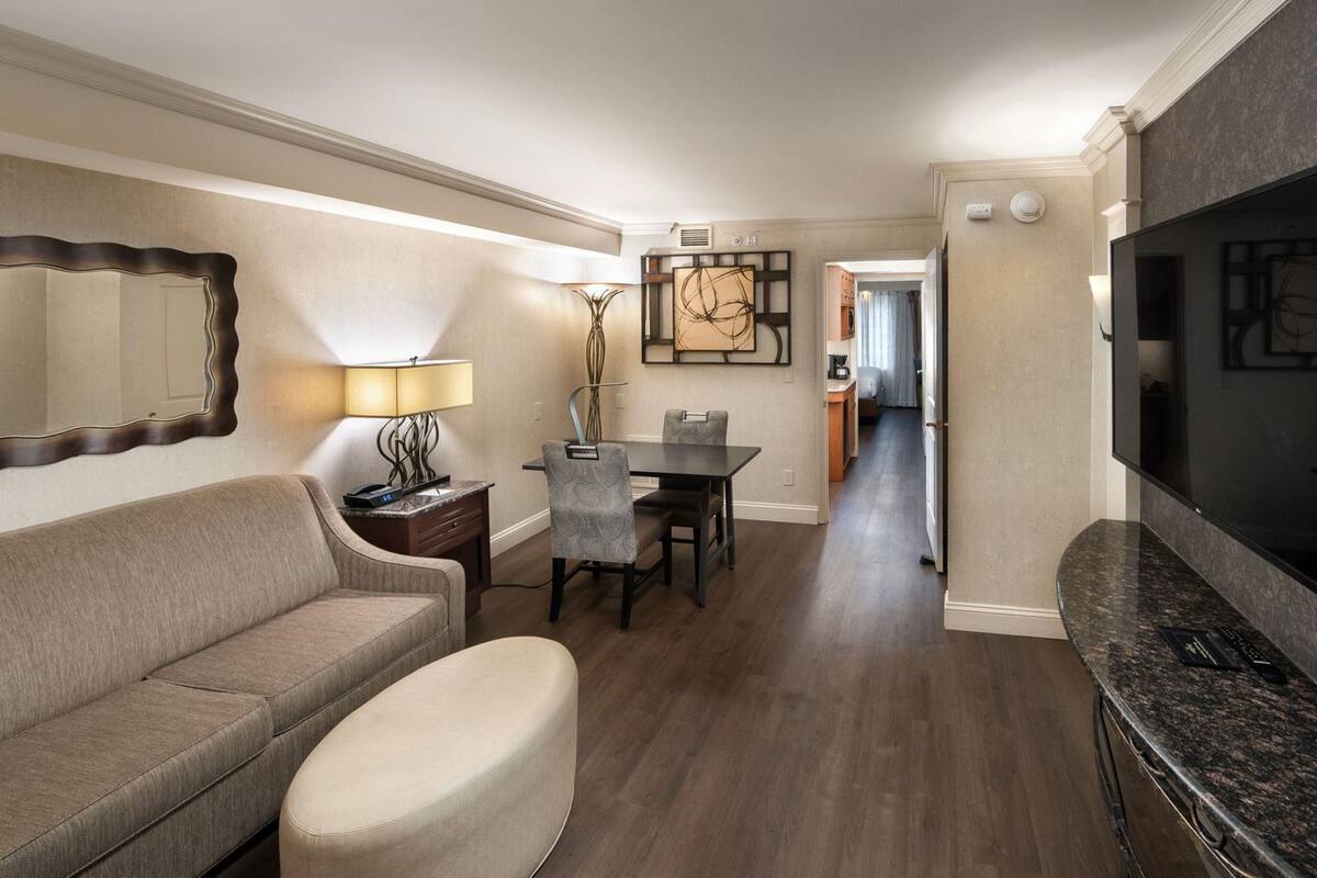 Una sala moderna con sofá, reposapiés, mesa de comedor y sillas. Hay un televisor a la derecha y un espejo grande a la izquierda.