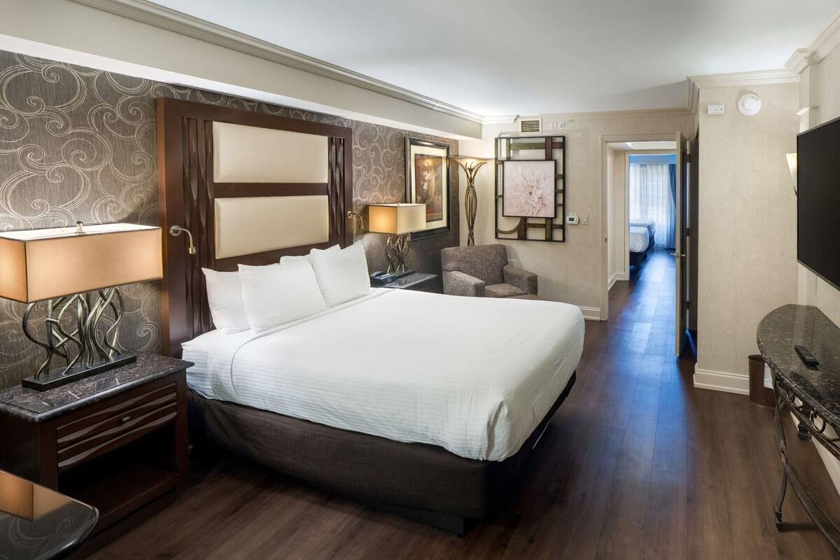 Habitación de hotel con una cama grande, un escritorio, una silla, un televisor y un pasillo que conduce a otra habitación.