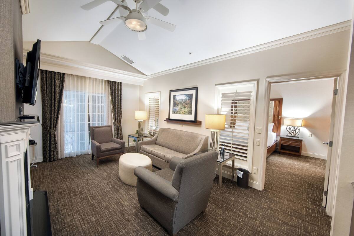 Habitación de hotel con sofá, sillón, reposapiés, ventilador de techo, dos lámparas, un televisor y un cuadro en la pared.