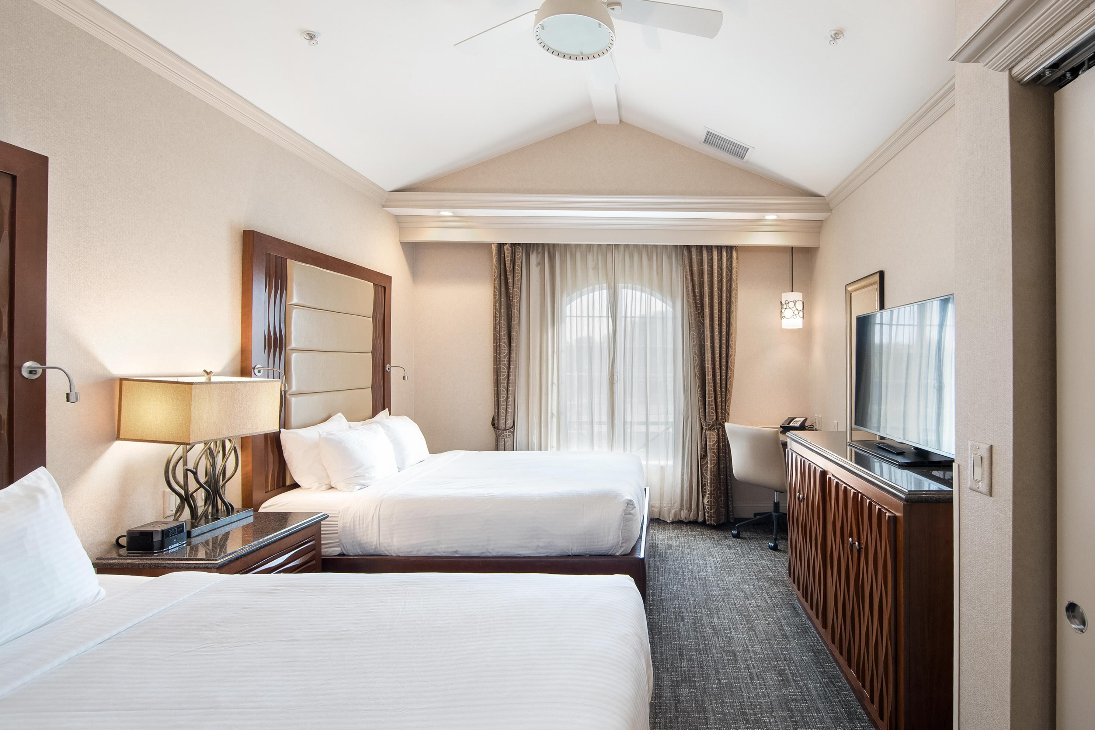 Quarto de hotel com duas camas, uma escrivaninha, uma televisão e um ventilador de teto.