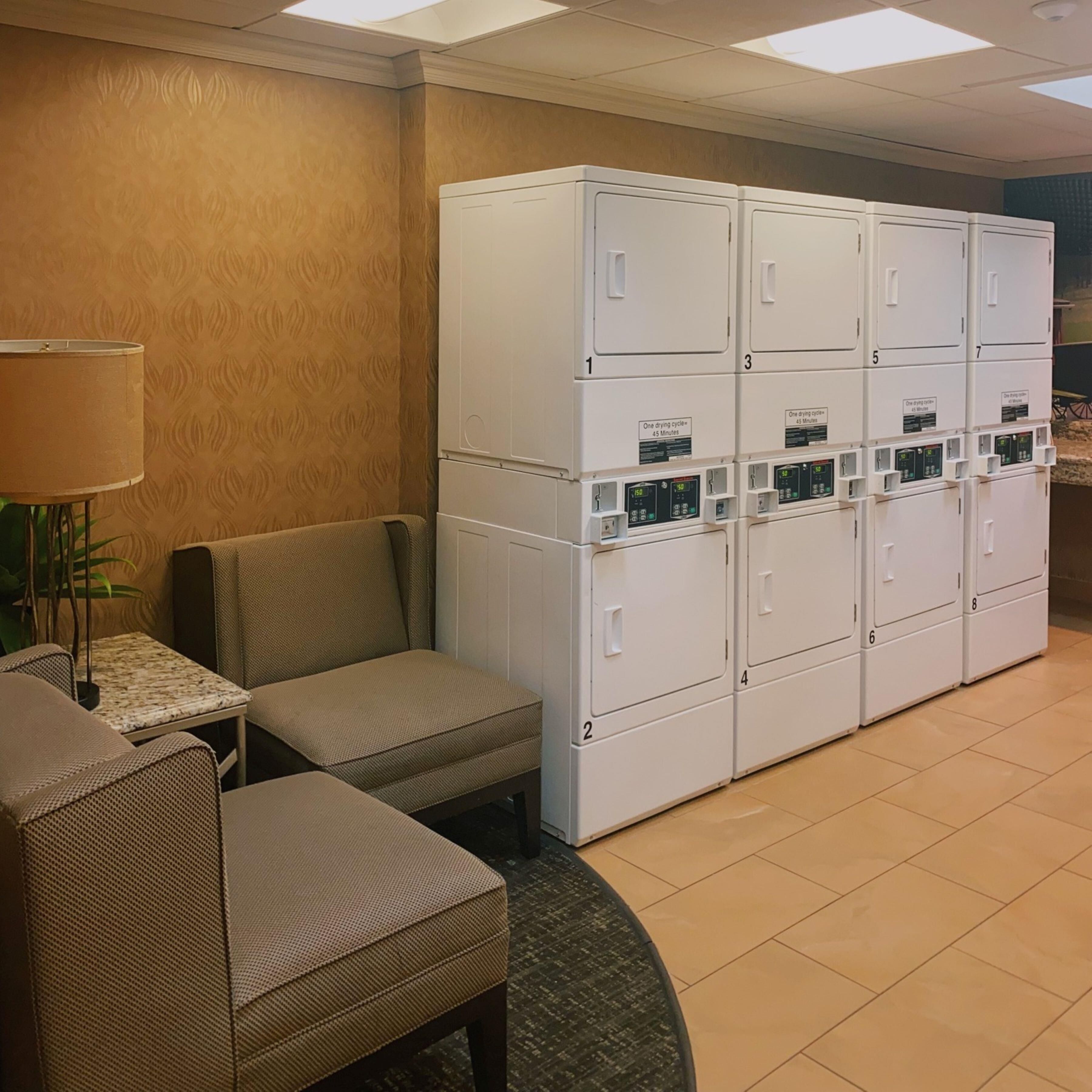 Quarto de hotel com área de estar, luminária e fileira de armários brancos com telas digitais.