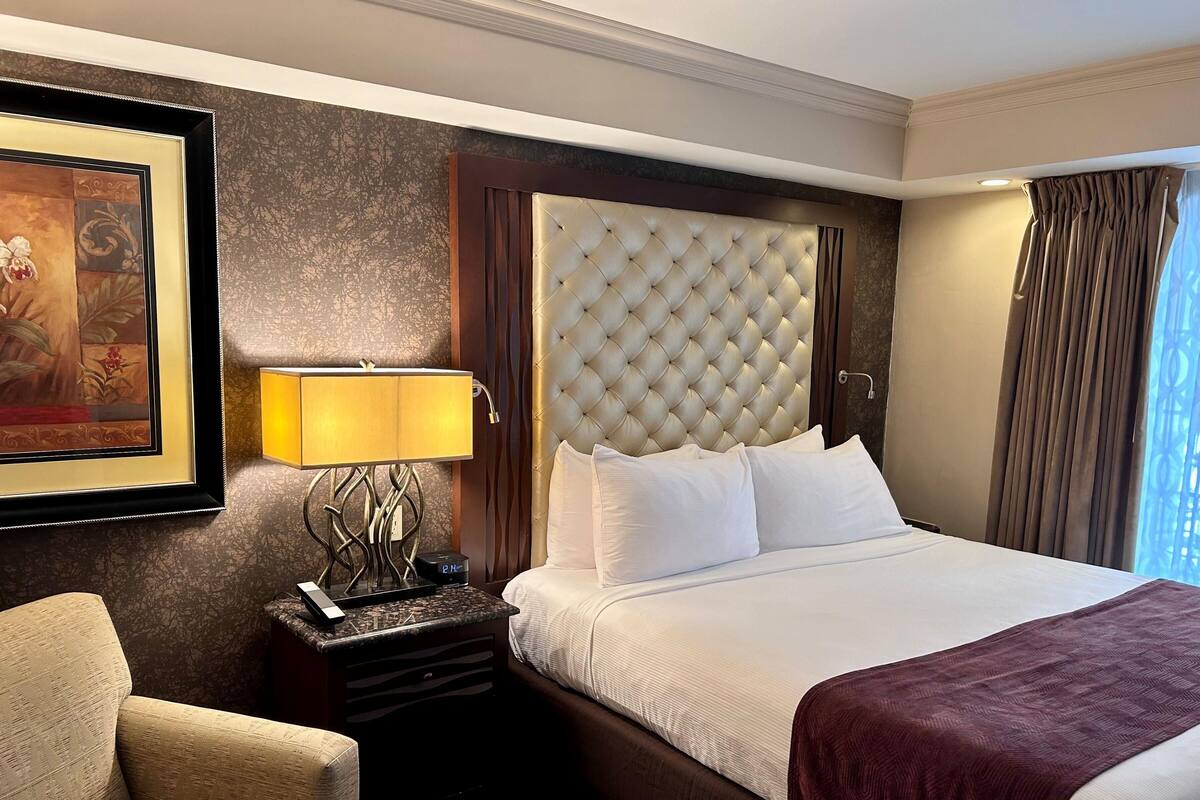 Una habitación de hotel con una cama grande, una mesita de noche con lámpara, una silla y un cuadro en la pared.