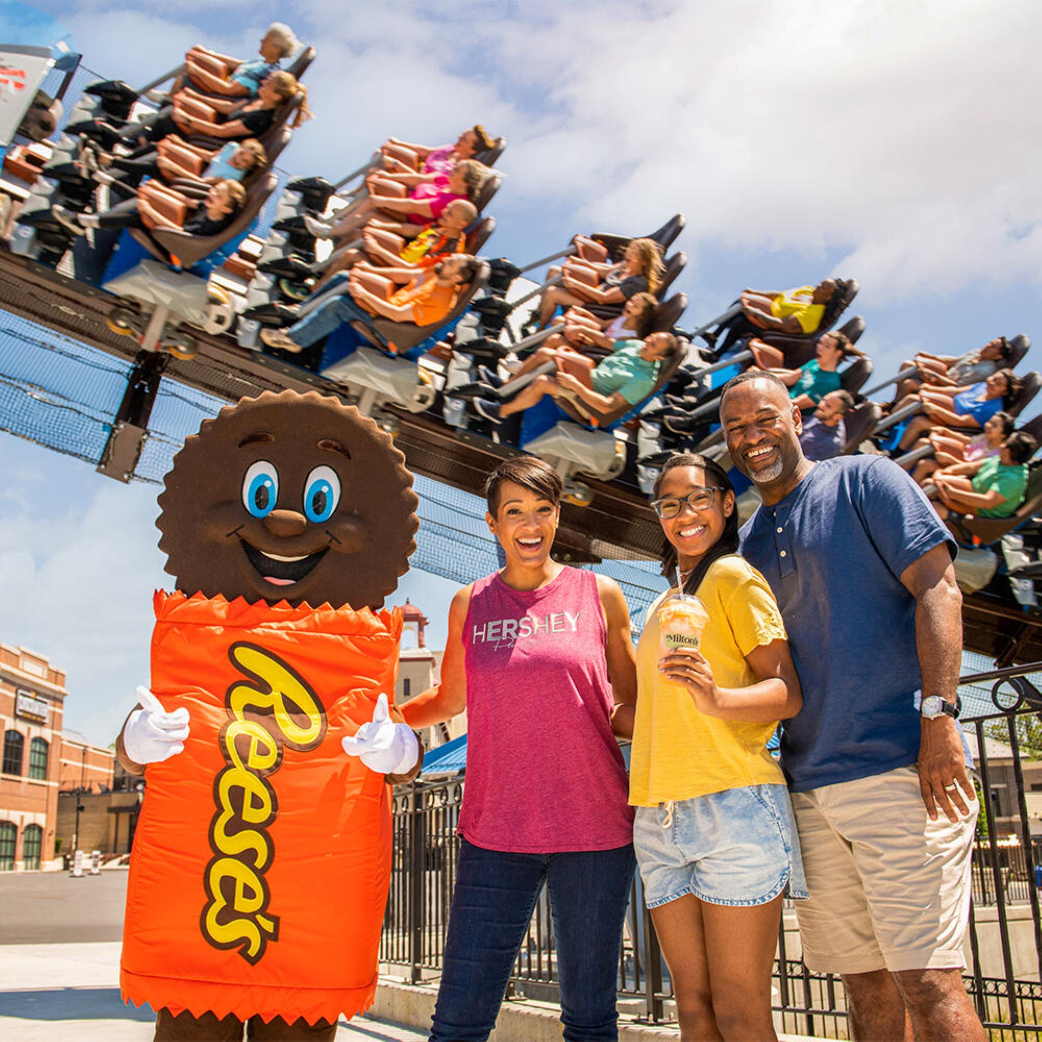A mascot in a costume standing in front of a roller coaster.
