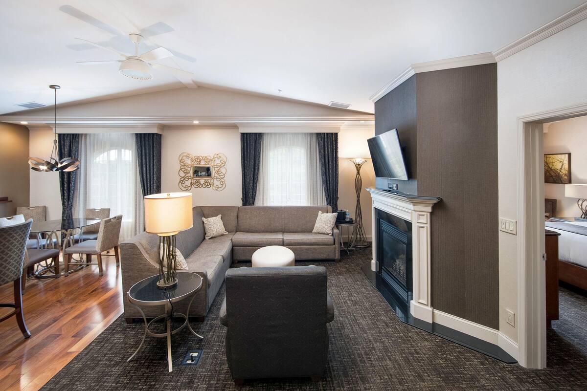 Un salón de hotel con sofá, otomán, mesa de comedor, sillas, chimenea y televisor.