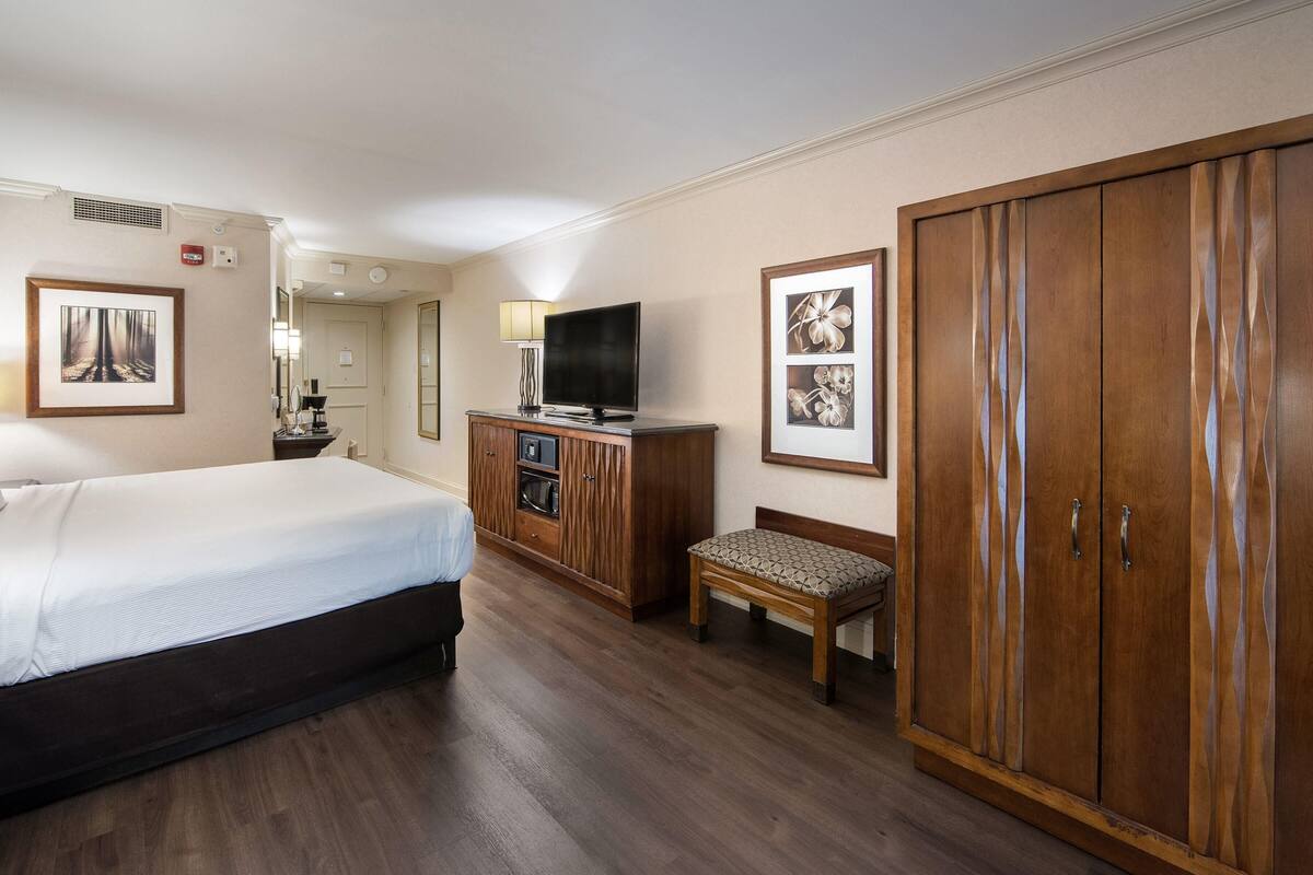 Habitación de hotel con cama, armario de madera, televisor, una mesita con lámpara y cuadros en la pared.