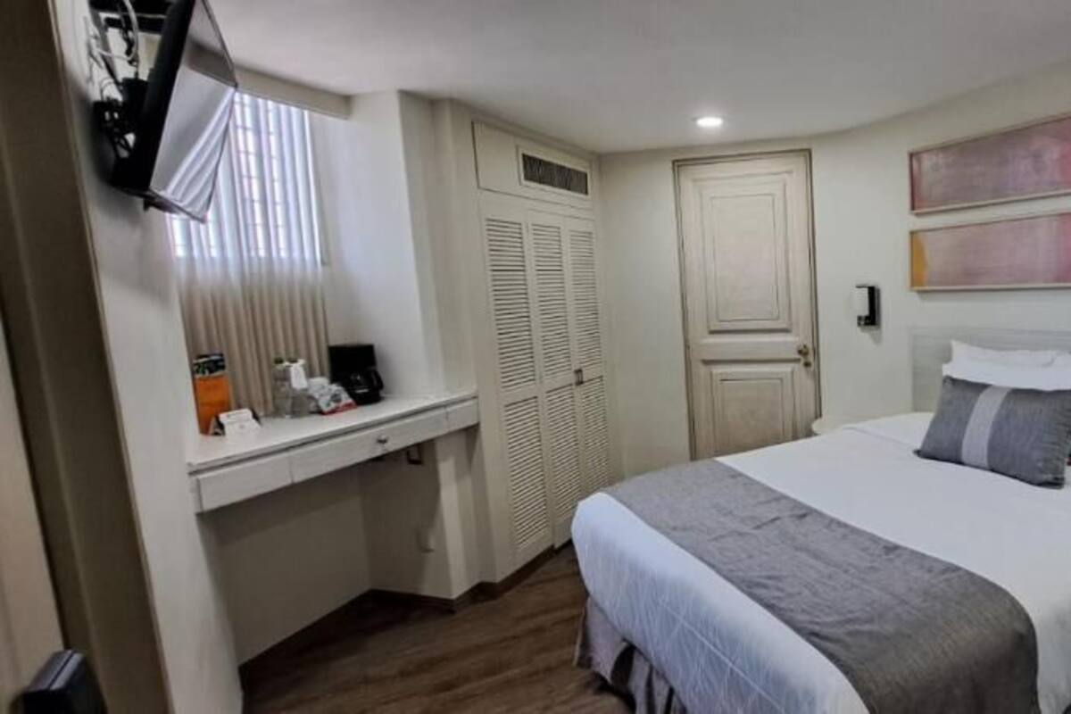 A hotel room with a bed, bedside table, lamp, and a window with blinds.