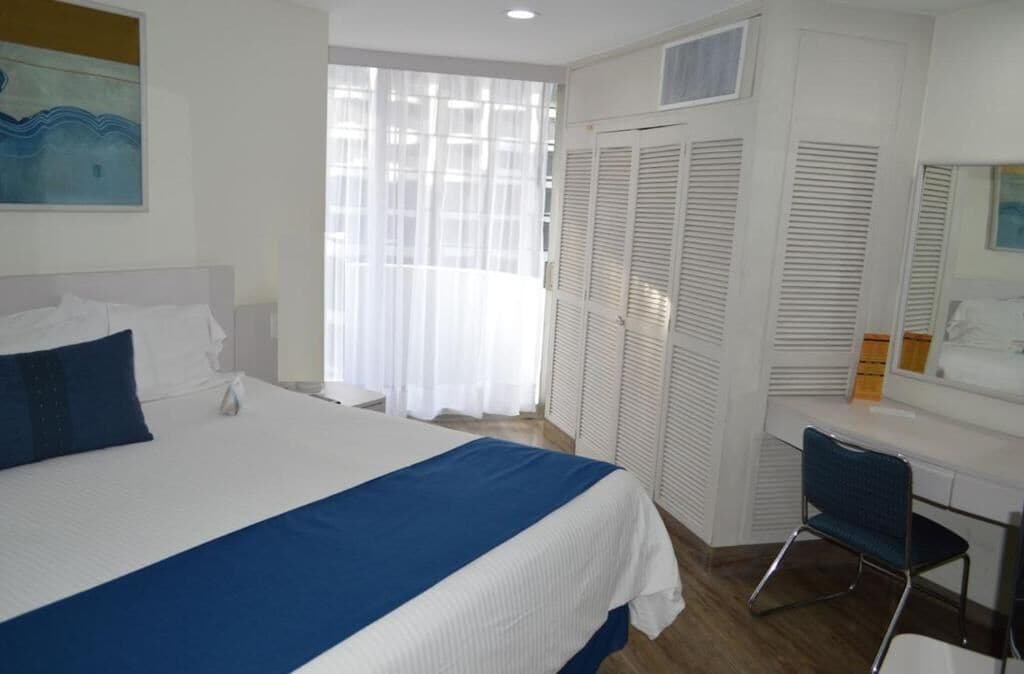 A hotel room with a large bed, a desk, and a chair. There is a window with curtains, a closet with louvered doors, and a painting on the wall.