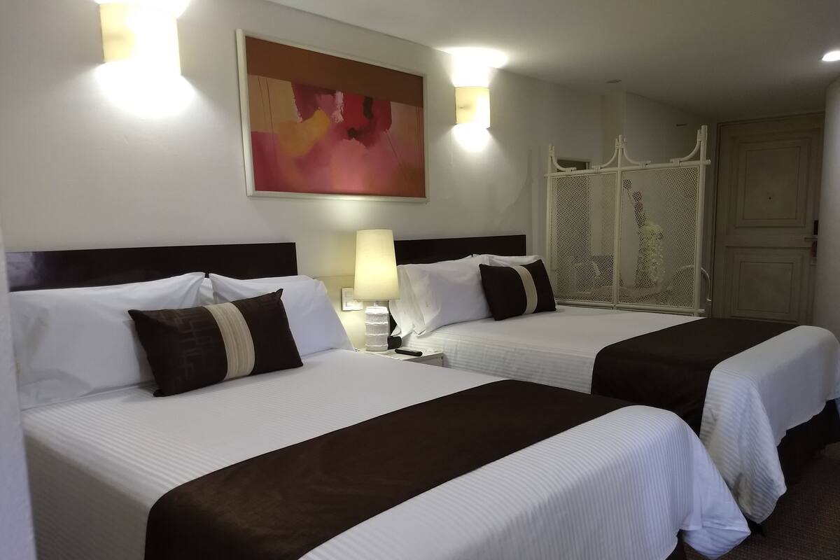 Une chambre d’hôtel avec deux lits, une table de chevet avec une lampe, une œuvre d’art accrochée au mur et une cloison blanche.