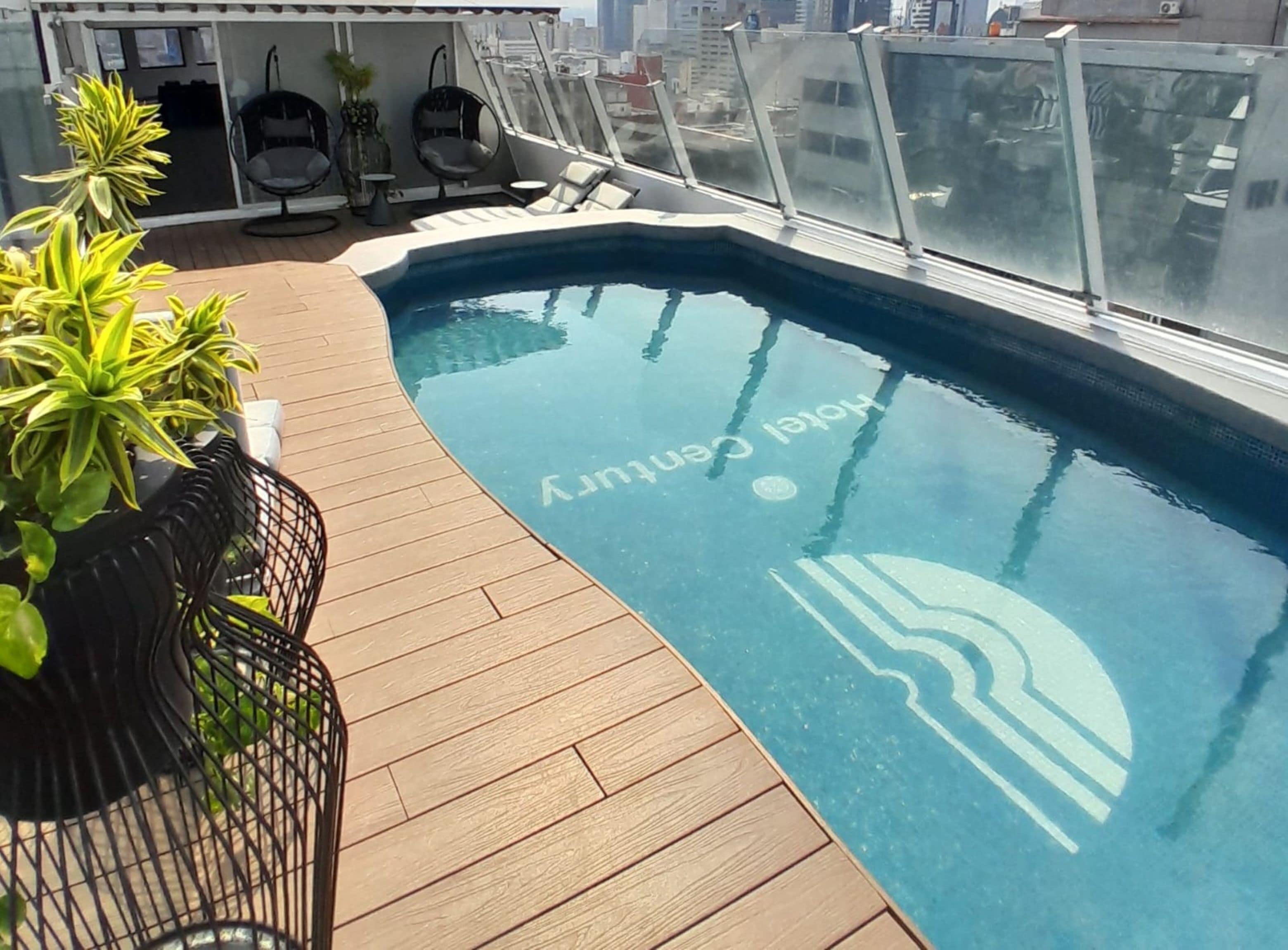 A rooftop pool with a wooden deck, potted plants, and a glass balustrade.