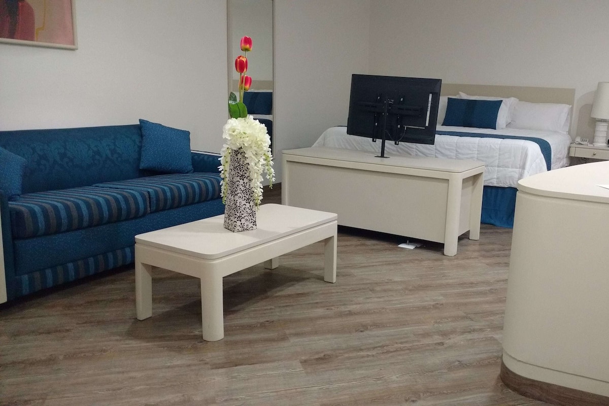 Une chambre d’hôtel avec un canapé bleu, une table blanche ornée d’un vase, un lit avec une literie blanche, un téléviseur posé sur un meuble et une table de chevet avec une lampe.