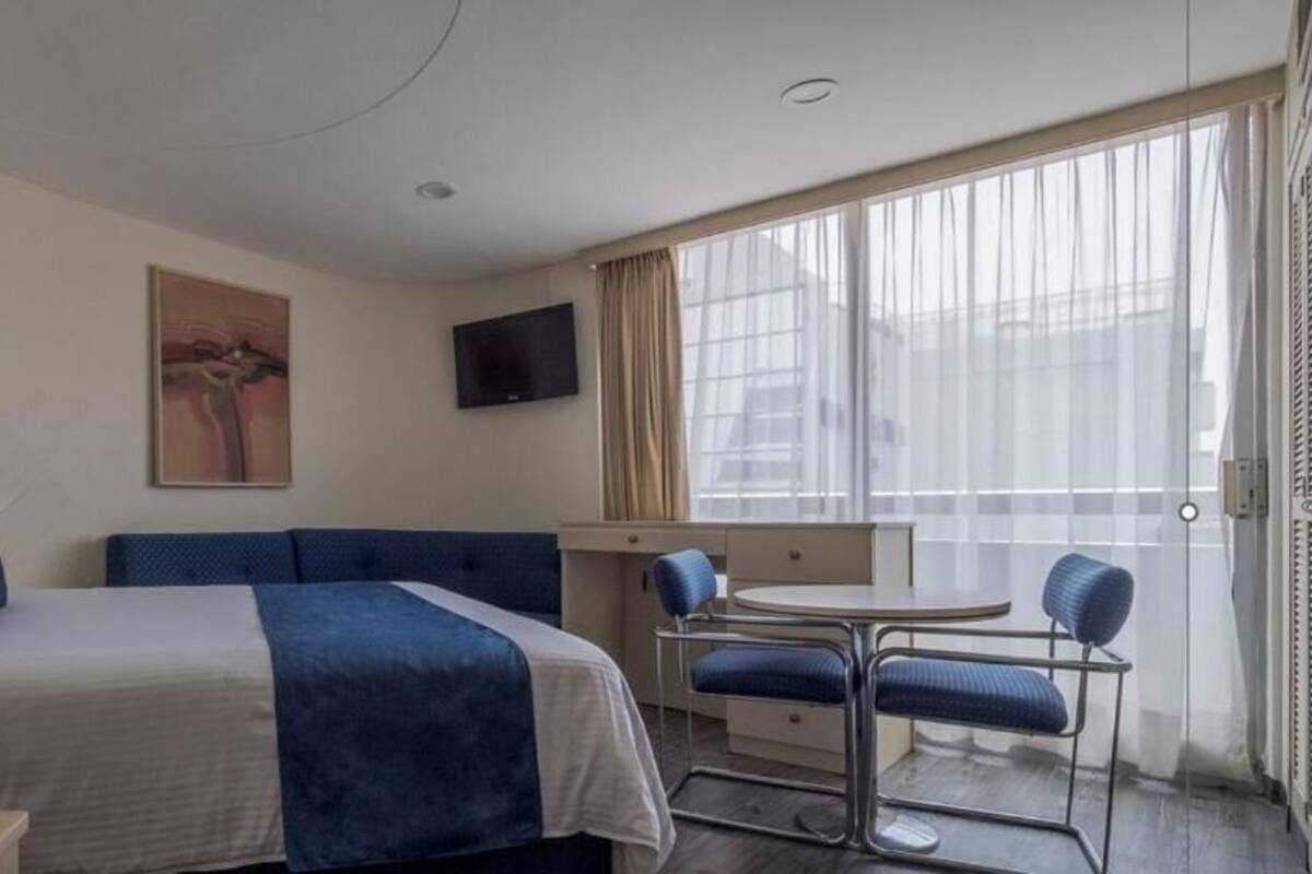 A hotel room with a bed, a small table with chairs, a TV mounted on the wall, and a large window with curtains.