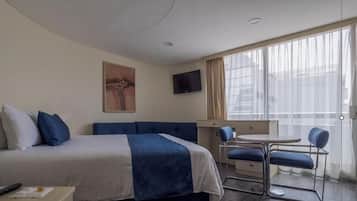 A hotel room with a bed, a small table with chairs, a TV mounted on the wall, and a large window with curtains.