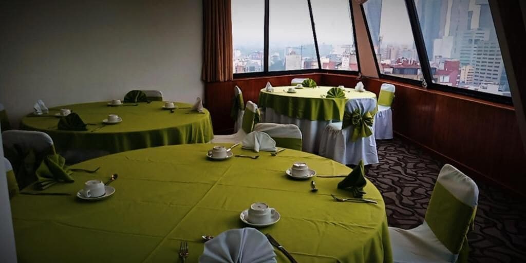 A dining area with round tables set for four, each with white tablecloths and green centerpieces. The room has large windows offering a city view.