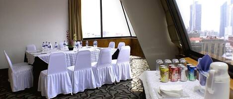 A conference room with a city view, equipped with a table set for meetings, white chairs, and a buffet table with snacks.