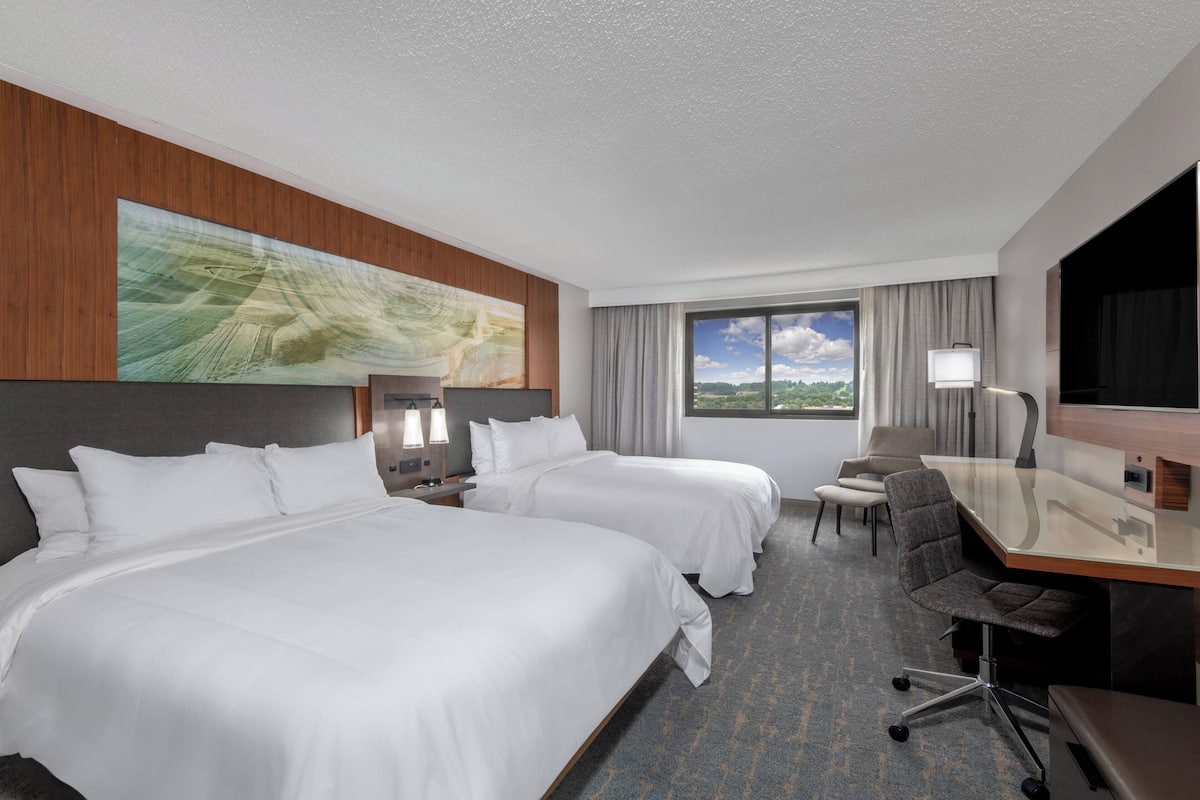 A hotel room with two beds, a desk, a chair, a TV, and a window with a view of the outdoors.