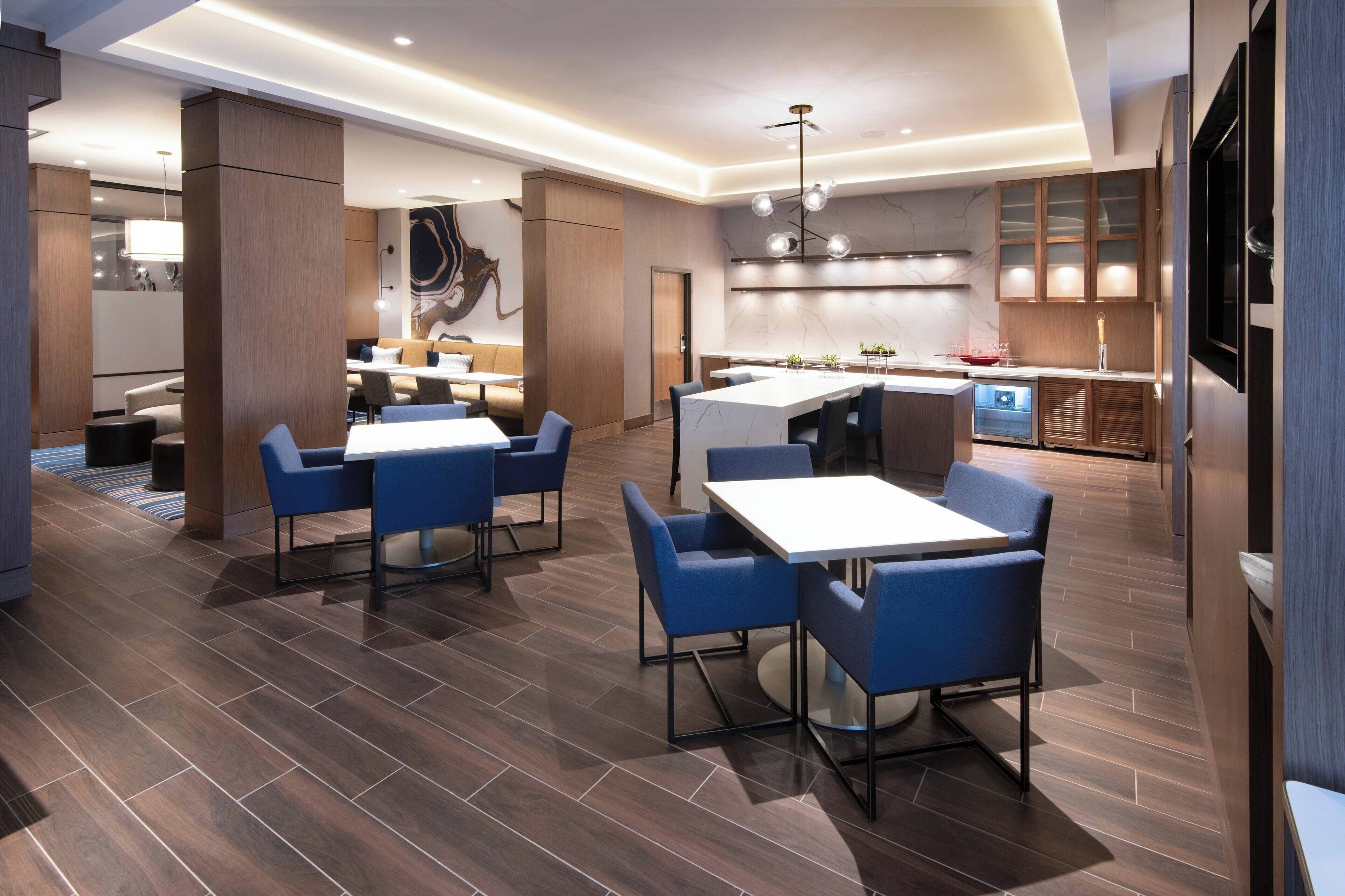 Un moderno vestíbulo de hotel con un amplio espacio de descanso, un bar central y una cocina visible al fondo.