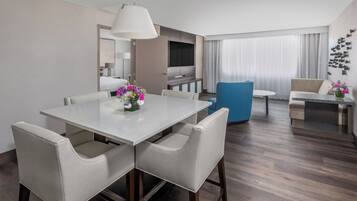 A modern hotel room with a dining area, a sofa, a TV, and a large window with curtains.