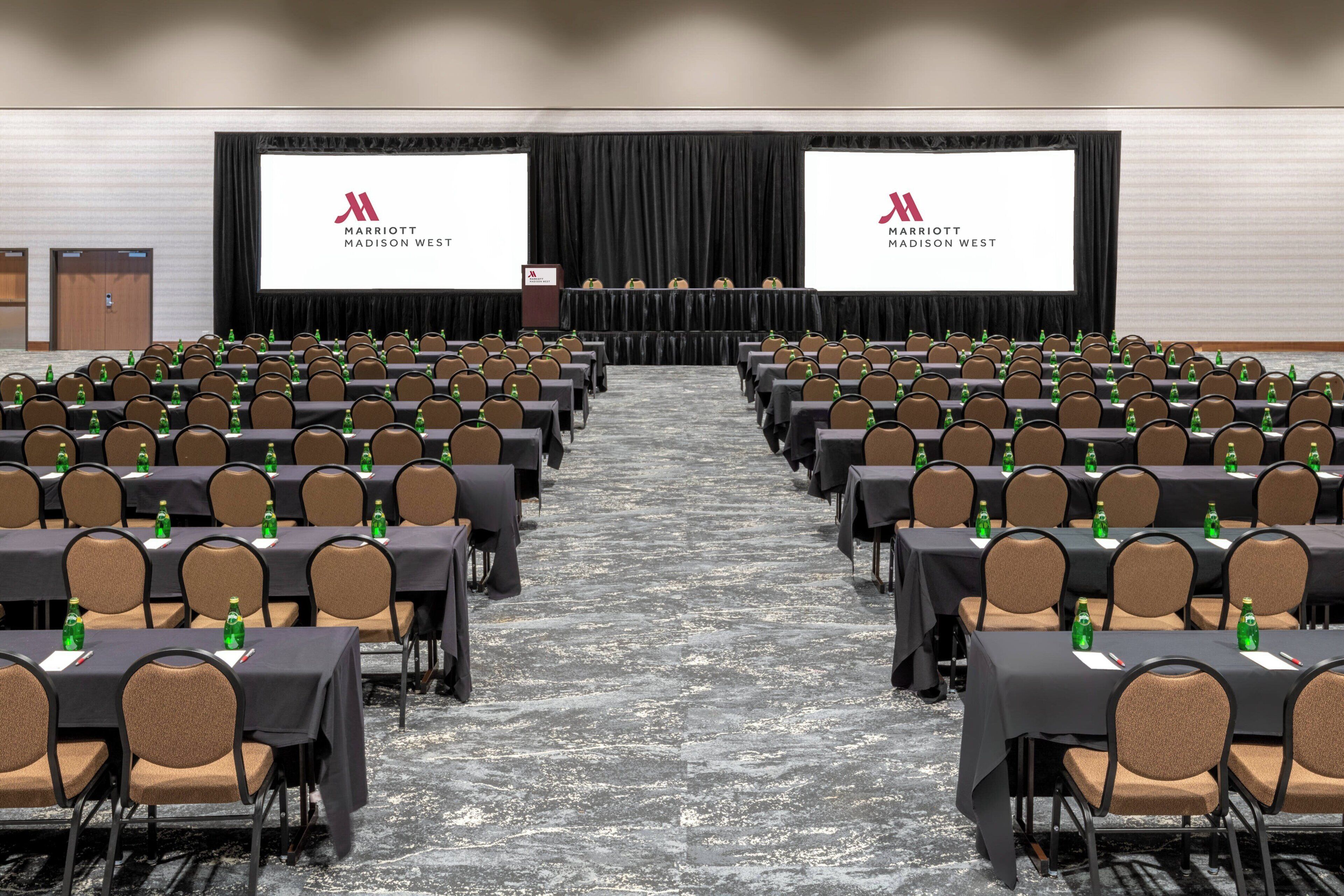 Un salón de conferencias con filas de mesas y sillas, un escenario con pantallas mostrando el logotipo de Marriott, y un piso alfombrado.