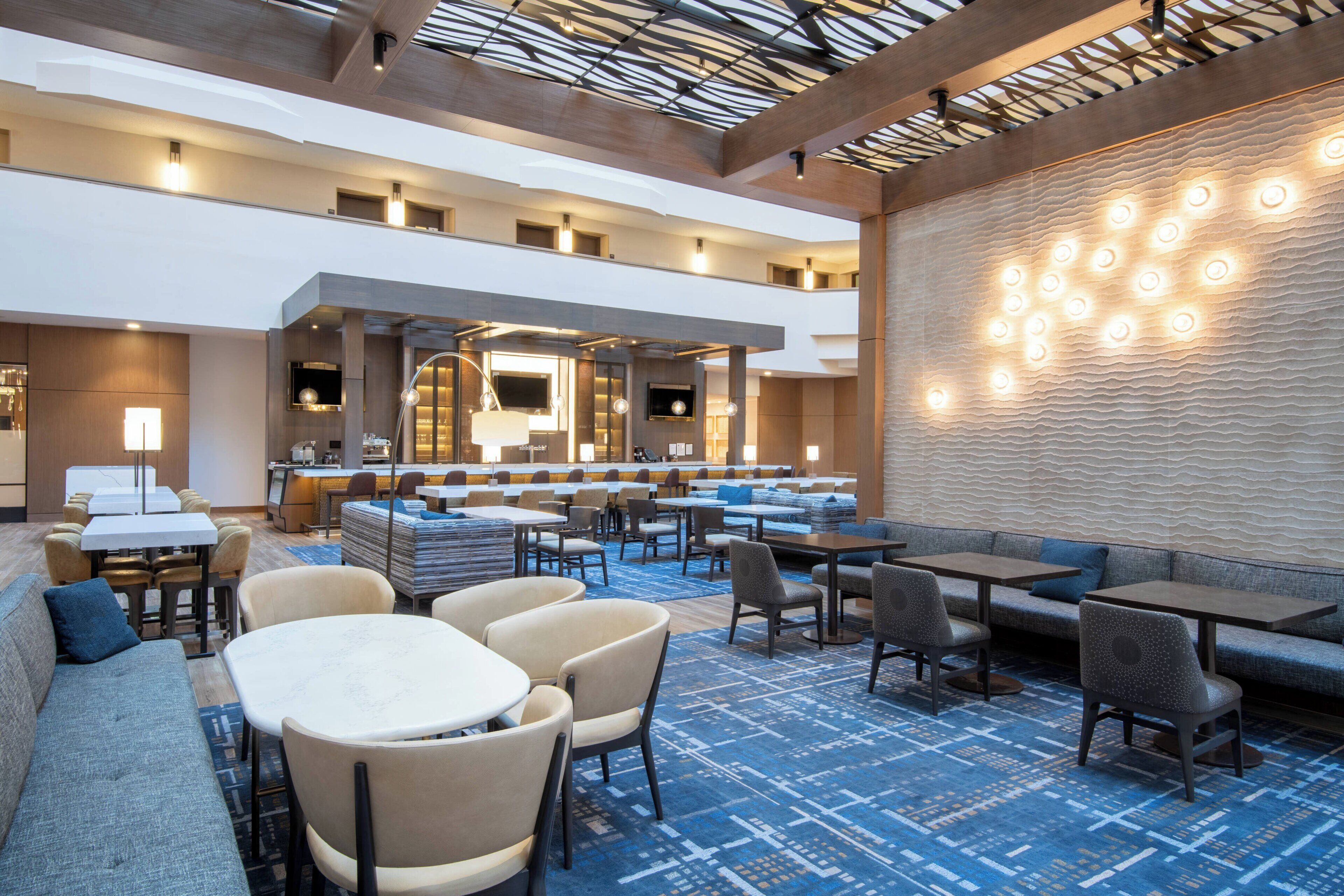 A hotel lobby with multiple seating areas, including sofas, tables, and chairs. There is a reception desk in the center with a counter and chairs. The lobby features a patterned wall and a ceiling with a grid-like design.
