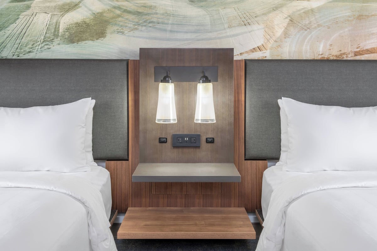 A hotel room with two beds, a wooden headboard, and a built-in shelf with two wall-mounted lights.
