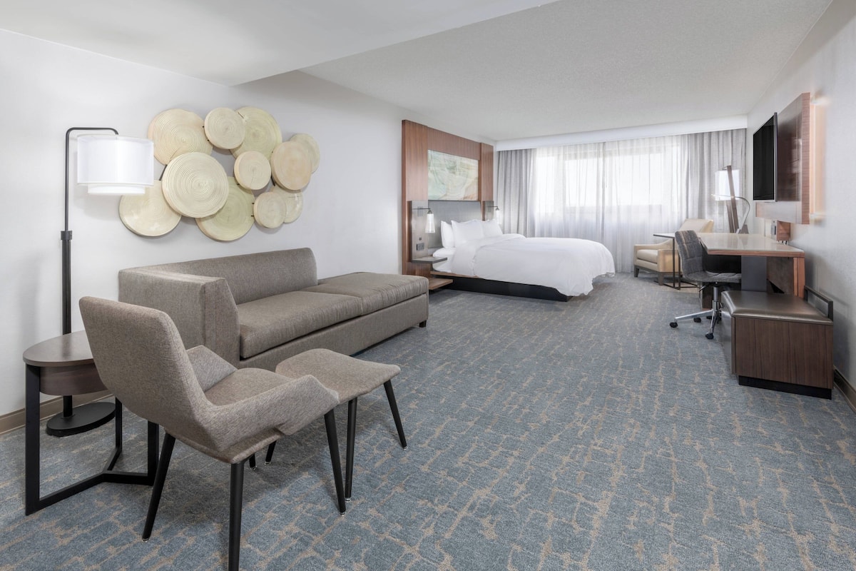 A hotel room with a bed, a sofa, a desk, a chair, a lamp, and a wall art piece.