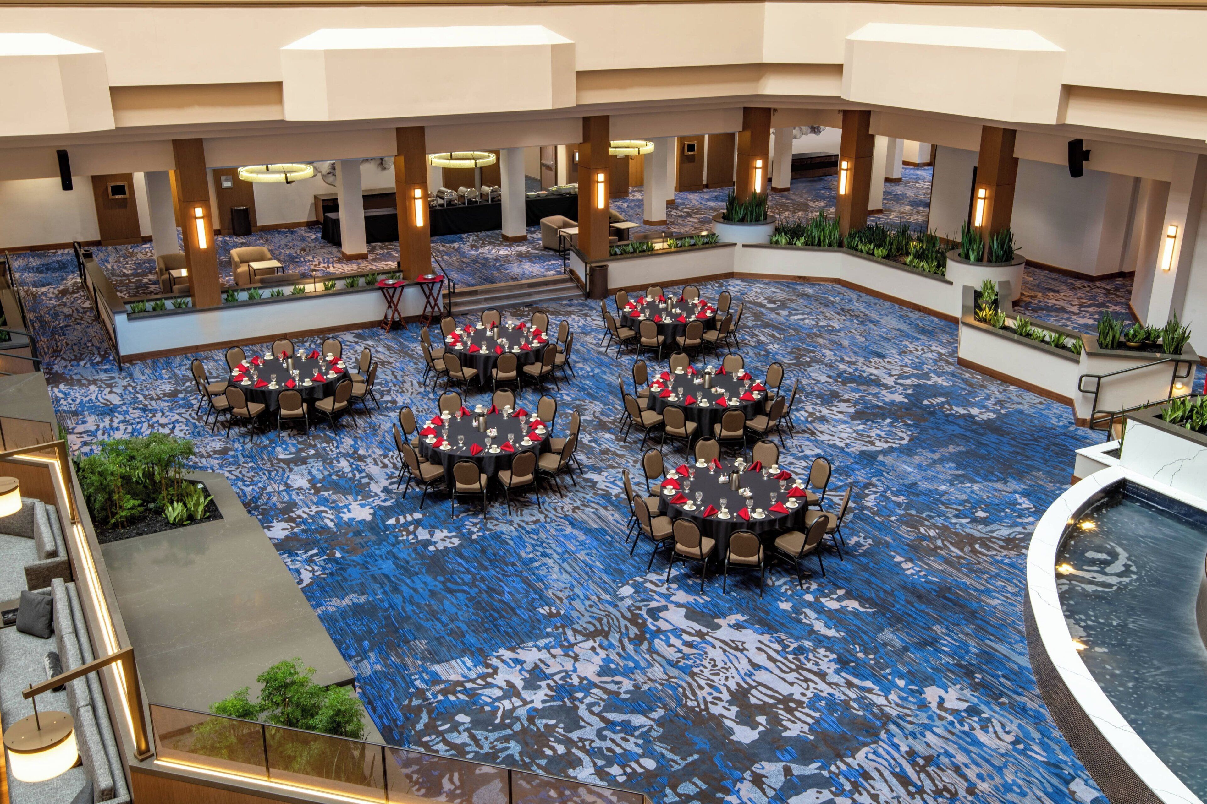 Un vestíbulo de hotel con varias mesas redondas dispuestas para comer, un área de piscina y un suelo cubierto de alfombra azul.