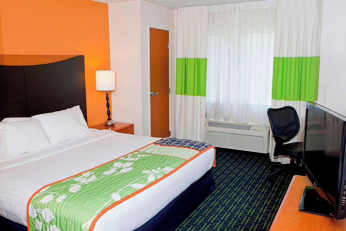 A hotel room with a bed, a desk with a computer, a chair, a lamp, and a window with curtains.