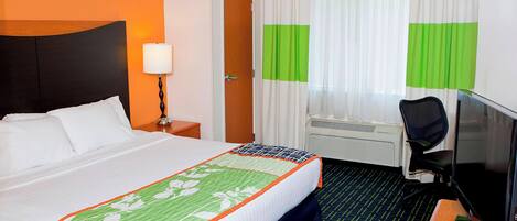 A hotel room with a bed, a desk with a computer, a chair, a lamp, and a window with curtains.
